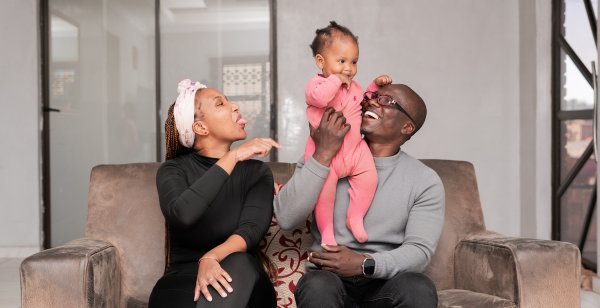 A joyful family smiles at home. Dad, Mom in black, and their 1-year-old daughter in pink.