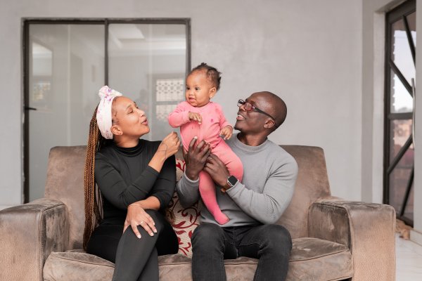 Black family smiles at home. Dad, Mom in black, and their 1-year-old daughter in pink.