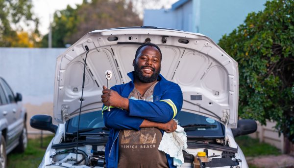 Black mechanic posing in front of a car for service repairs on engine wearing blue uniform.