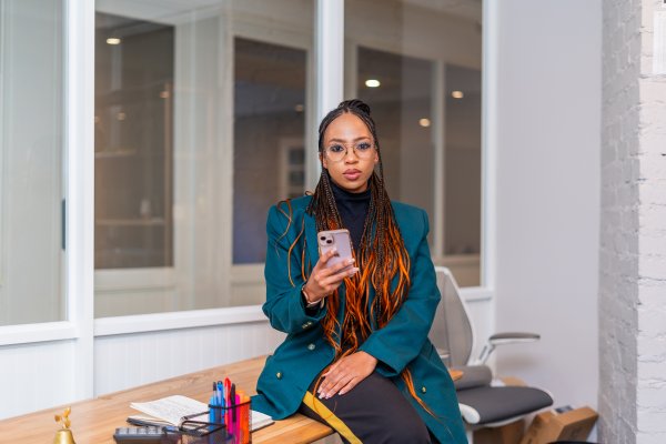 Black woman sitting on desk at work holding a phone and looking at the camera.