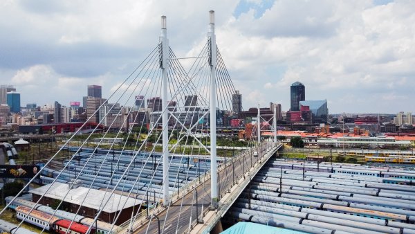 Close up drone photo of Nelson Mandela bridge from above with Johannesburg cbd in the background and trains under the bridge.