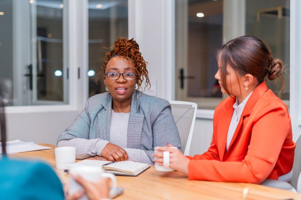 Mature black woman sitting at desk wearing glasses speaking to a candidate at an interview at the office for employment.