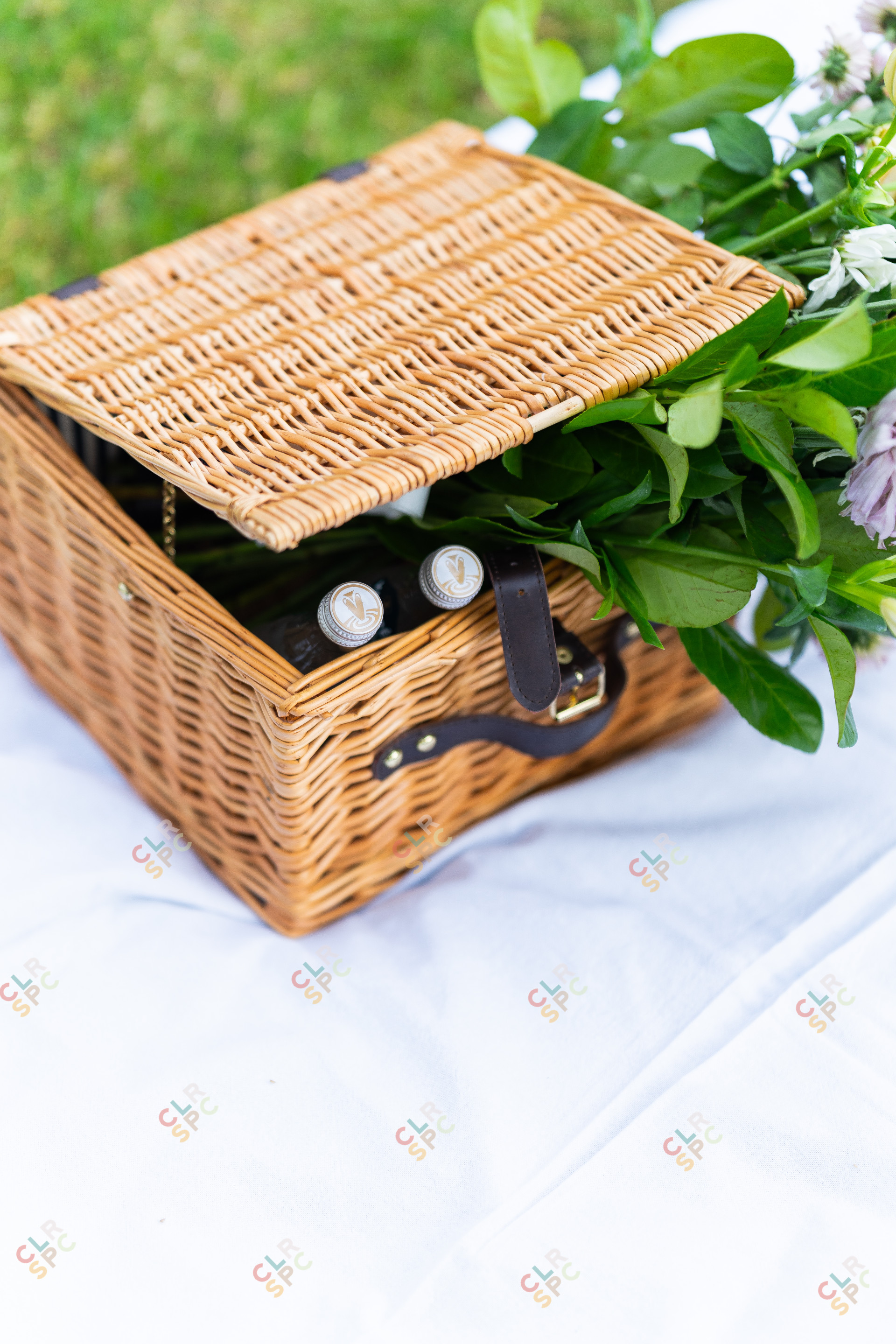 Picnic basket with vamper water and flowers inside on a white sheet.