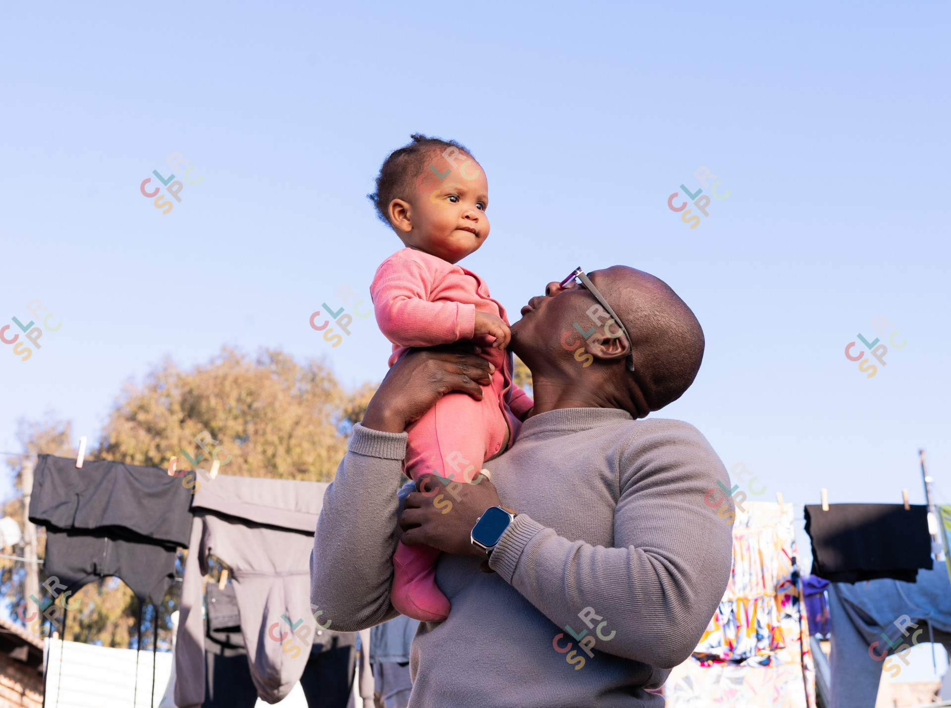 Black father holding daughter wearing pink outside with clothes in the background.